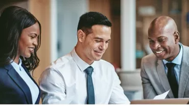 Three people in formal attire smiling at a laptop.