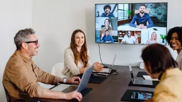 Happy employees sitting in an office conference room video calling remote teams on a large screen