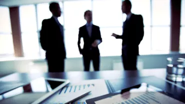 Three people in a meeting room with documents and graphs on a table.