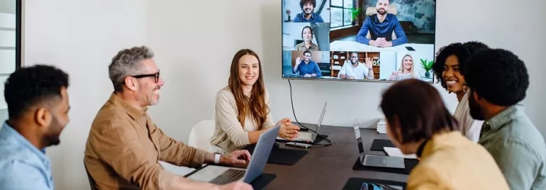 Happy employees sitting in an office conference room video calling remote teams on a large screen